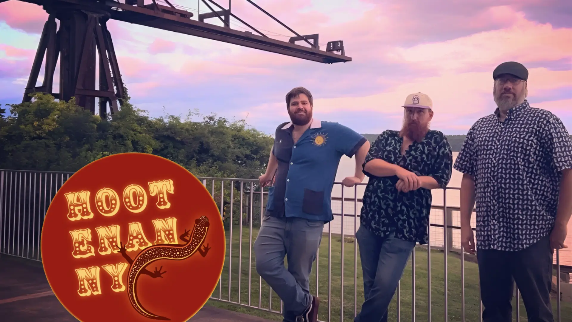 Three men pose in front of a railing and an industrial structure, with a circular "HOOTENANNY" logo,